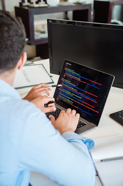The image depicts a person working at a desk with a laptop displaying lines of colorful code. The individual, wearing a light blue shirt, is seated in front of a computer monitor, which is turned off,