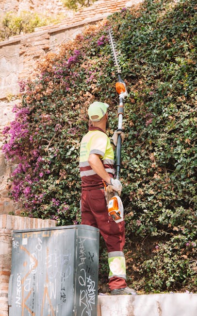 A gardener wearing a pale green cap, yellow and maroon high-visibility vest, and maroon work trousers is trimming a dense hedge with a long-reach pruning tool in a garden. The hedge, which has a mix o